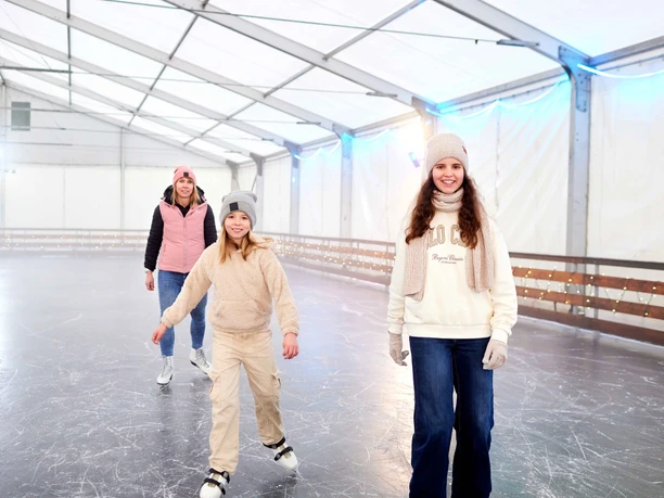 Drei Personen lächeln beim Schlittschuhlaufen in einer überdachten Eisbahn.