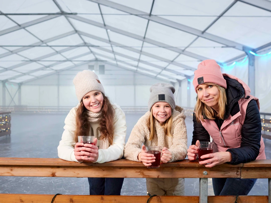 Eisbahn in Königstein Drei Frauen in Winterkleidung lehnen an einer Brüstung und halten Becher.