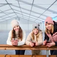 Eisbahn in Königstein Drei Frauen in Winterkleidung lehnen an einer Brüstung und halten Becher.