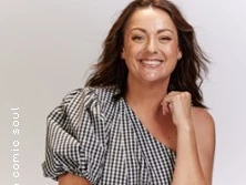 Lächelnde Frau mit kariertem Oberteil vor hellem Hintergrund, locker posierend.