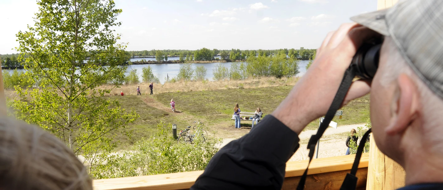Vogelbeobachtungsturm am Bargerveen in Twist ©Naturpark Moor-Veenland (29).jpg Mann beobachtet mit Fernglas vom Holzturm Menschen und Landschaft am See im Naturpark Moor-Veenland
