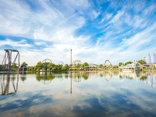 heide-park-resort-soltau-seeblick.jpg Achterbahnen und Aussichtsturm des Heide Park Resort spiegeln sich im ruhigen See unter blauem Himmel.