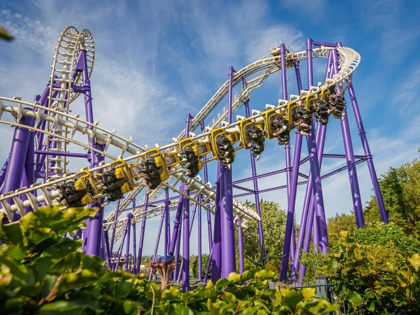 heide-park-resort-soltau-toxic-garden.jpg Achterbahn mit gelb-violetten Schienen fährt in einer steilen Kurve über grüne Büsche hinweg.