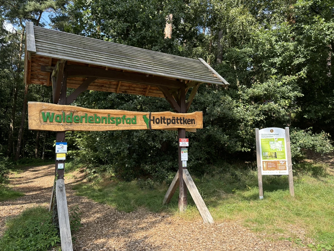 Frerener Holtpättken ©Emsland Tourismus GmbH (1).JPEG Holztor mit Schild „Walderlebnispfad Holtpättken“ am Waldrand, umgeben von dichtem Grün und Infotafel.