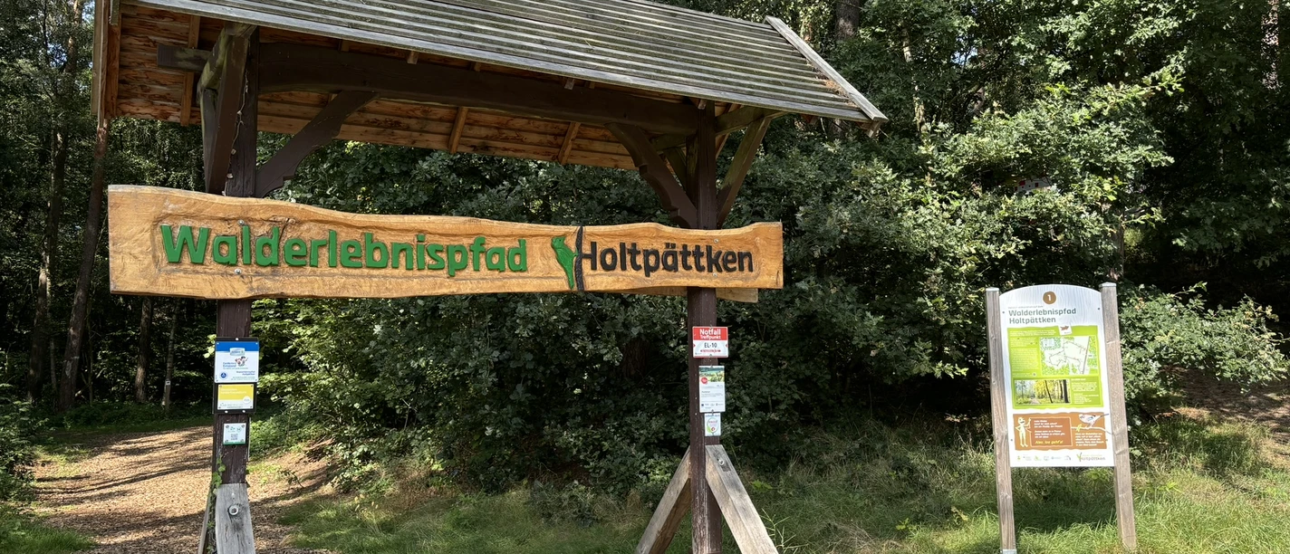 Frerener Holtpättken ©Emsland Tourismus GmbH (1).JPEG Holztor mit Schild „Walderlebnispfad Holtpättken“ am Waldrand, umgeben von dichtem Grün und Infotafel.
