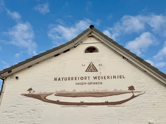 Naturresort Weserinsel Weißer Gebäudegiebel mit Schriftzug Naturresort Weserinsel und gemalter Flusslandschaft.