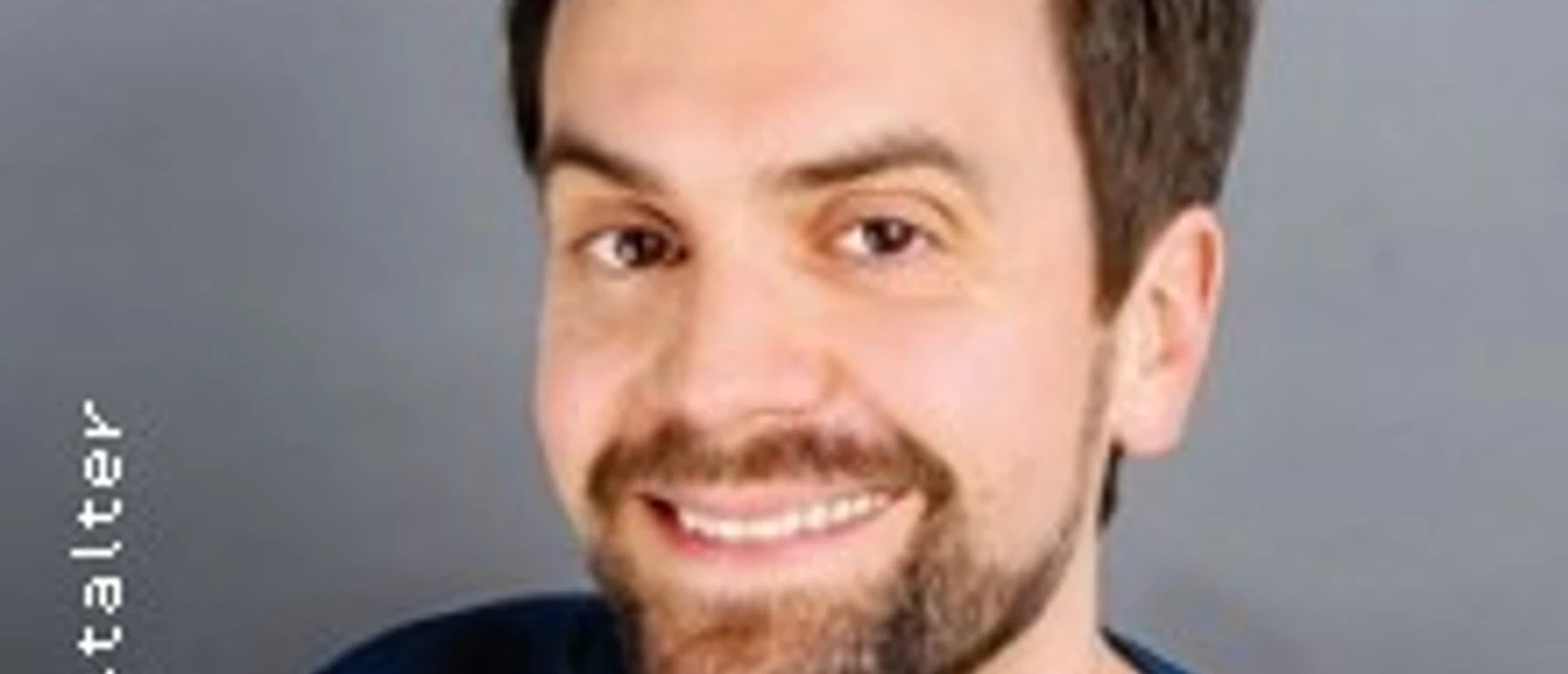 Ein Mann mit Bart lächelt vor neutralem Hintergrund.