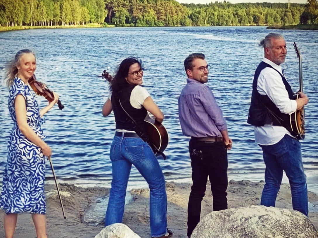 1 Band to Chill.jpg Vier Musiker mit Geige und Gitarren stehen am Ufer eines Sees und blicken lächelnd zur Kamera.