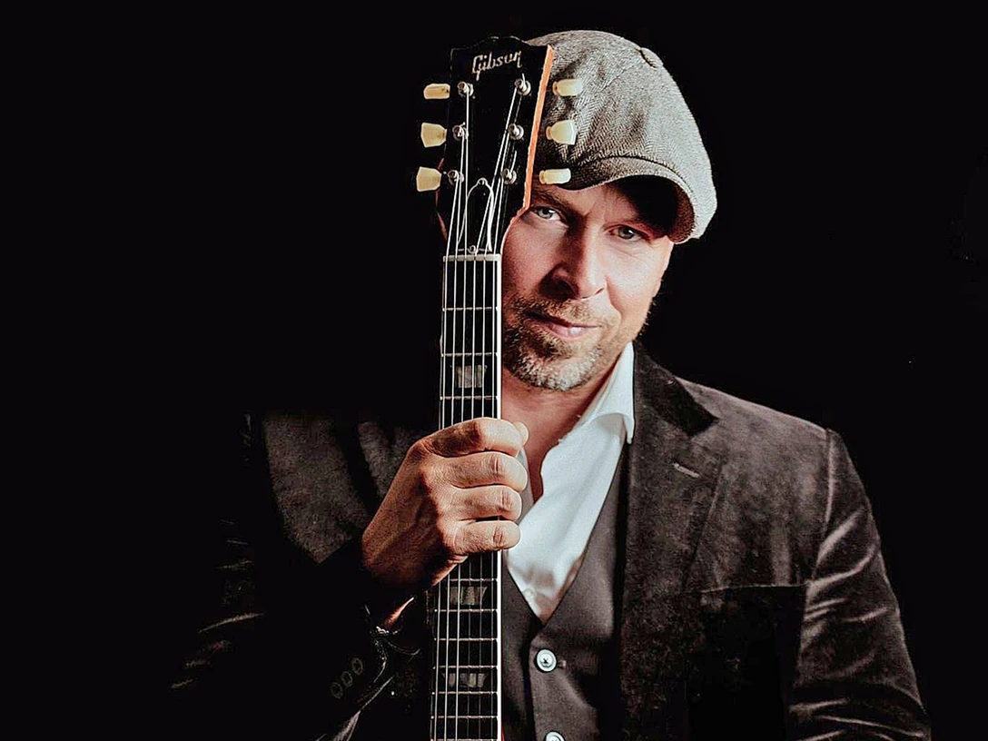 2 Oliver Jüchems.jpg Mann mit Schiebermütze hält eine Gitarre vor sich, stilvolles Porträt vor dunklem Hintergrund.