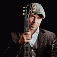 Oliver Jüchems.jpg Mann mit Schiebermütze hält eine Gitarre vor sich, stilvolles Porträt vor dunklem Hintergrund.