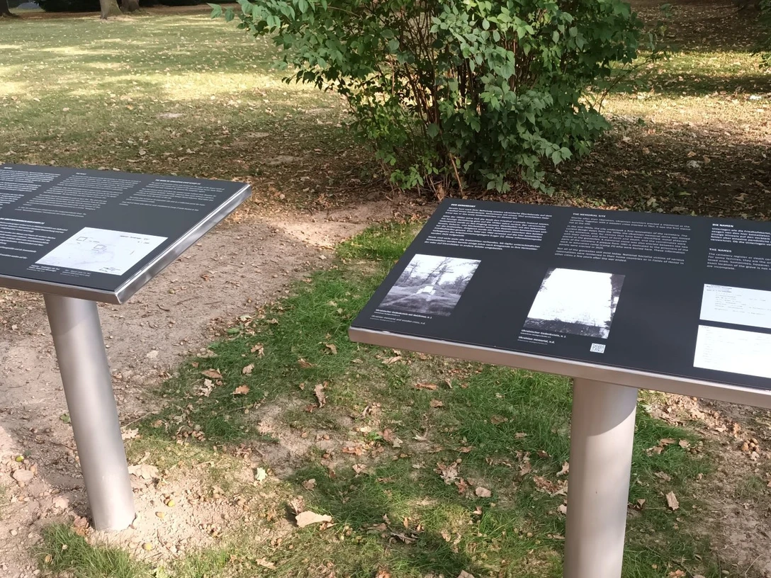 Ehrenfriedhof_Westerholz (3)_HD_16zu9.jpg Zwei Informationstafeln mit Texten und Fotos auf einem Ehrenfriedhof, umgeben von Grün und Bäumen.