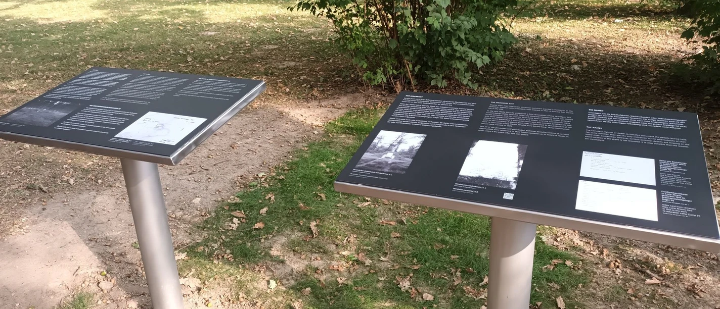 Ehrenfriedhof_Westerholz (3)_HD_16zu9.jpg Zwei Informationstafeln mit Texten und Fotos auf einem Ehrenfriedhof, umgeben von Grün und Bäumen.