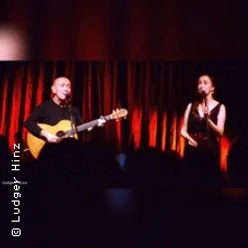 Zwei Künstler auf Bühne, einer mit Gitarre, beide im warmen Scheinwerferlicht vor rotem Vorhang.