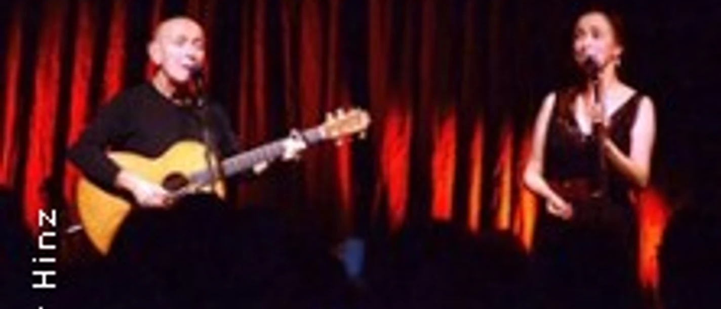 Zwei Künstler auf Bühne, einer mit Gitarre, beide im warmen Scheinwerferlicht vor rotem Vorhang.