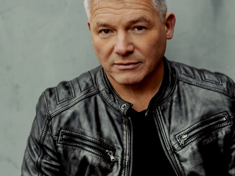 Porträt von Andreas Winkelmann Person mit grauen Haaren in dunkler Lederjacke vor grauer Wand, direkter, ruhiger Blick in Kamera