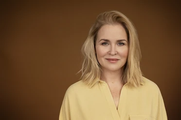 Judith Hoersch Frau mit blondem Haar und hellem Hemd vor braunem Hintergrund, freundlich und ruhig blickend.