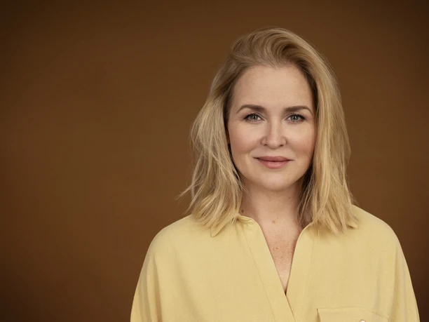 Judith Hoersch Frau mit blondem Haar und hellem Hemd vor braunem Hintergrund, freundlich und ruhig blickend.