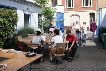 ehrenfeldstudios Personen in einem sonnigen Innenhof an Tischen im Gespräch und beim Vorbereiten einer Aktivität
