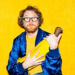 Mann mit Brille hält ein übergroßes gelbes Objekt vor einer leuchtend gelben Wand.
