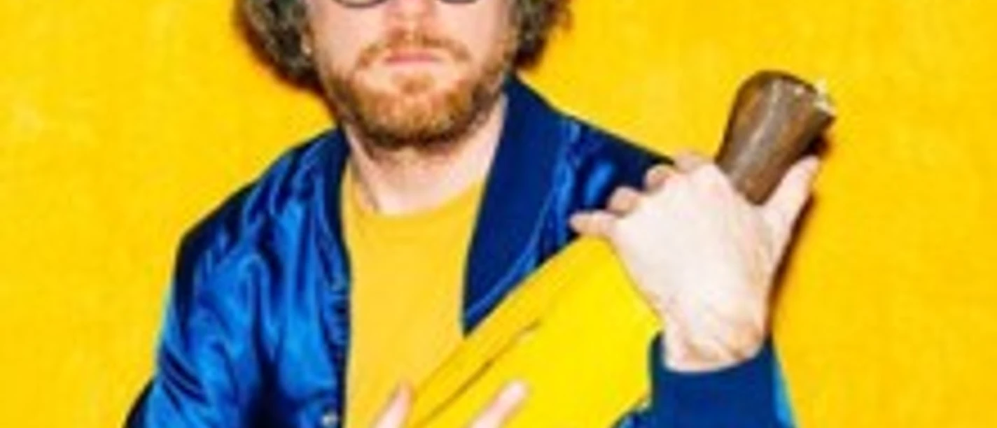 Mann mit Brille hält ein übergroßes gelbes Objekt vor einer leuchtend gelben Wand.