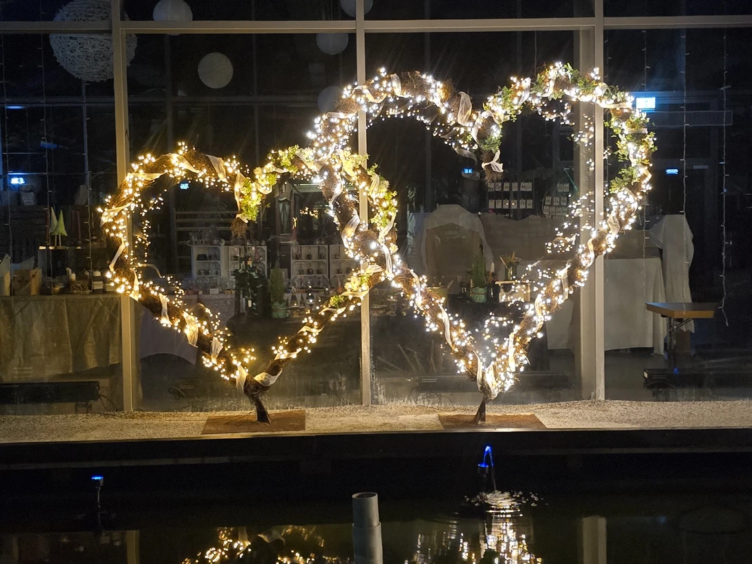 1000194320.jpg Zwei leuchtende Herzskulpturen aus Lichterketten spiegeln sich nachts im Wasser vor einer Glasfassade.