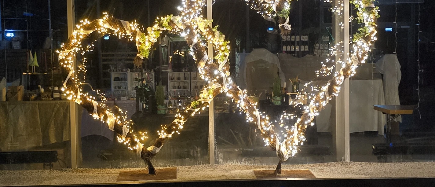 1000194320.jpg Zwei leuchtende Herzskulpturen aus Lichterketten spiegeln sich nachts im Wasser vor einer Glasfassade.