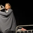 Windsbraut_Theater Rosenfisch (c) Lea Schornstein.jpg Eine Frau sitzt mit einer Decke umschlungen auf einem Patientenbett und hält eine Figur in der Hand.