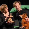 Eine Frau und ein Man sitzen beide schwarz gekleidet auf einer Theaterbühne und halten die Figur von einem Hasen und einem Fuchs in den Händen.