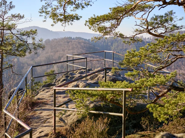 Aussichtspunkt mit Geländer auf einem Felsen, umgeben von Bäumen, Blick auf Berge.