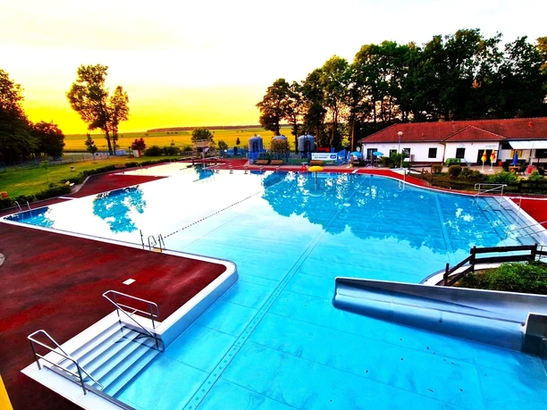 Overview of the Böhlitz outdoor pool complex with swimming pool, adjacent sunbathing lawn, trees and buildings in the background.
