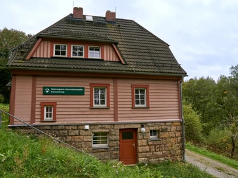 Beizehaus Nationalpark Infostelle Hinterhermsdorf in der Waldhusche Zweistöckiges Haus mit rosa Fassade und Steinfundament in ländlicher Umgebung.