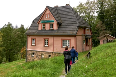 Drei Personen wandern auf einem Pfad zu einem rosa Haus im Wald.