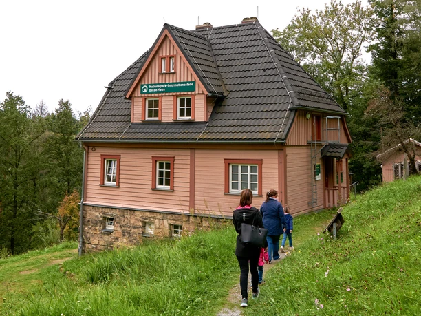 Drei Personen wandern auf einem Pfad zu einem rosa Haus im Wald.