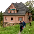Beizehaus Nationalpark Infostelle Hinterhermsdorf in der Waldhusche Drei Personen wandern auf einem Pfad zu einem rosa Haus im Wald.