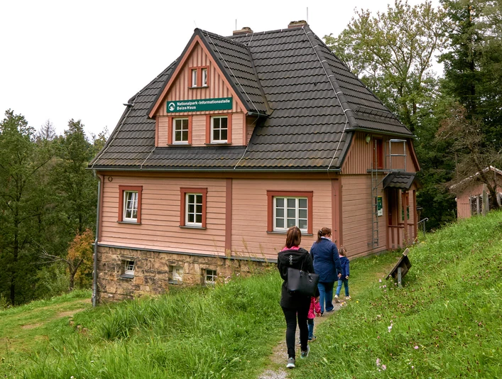 Beizehaus Nationalpark Infostelle Hinterhermsdorf in der Waldhusche Drei Personen wandern auf einem Pfad zu einem rosa Haus im Wald.