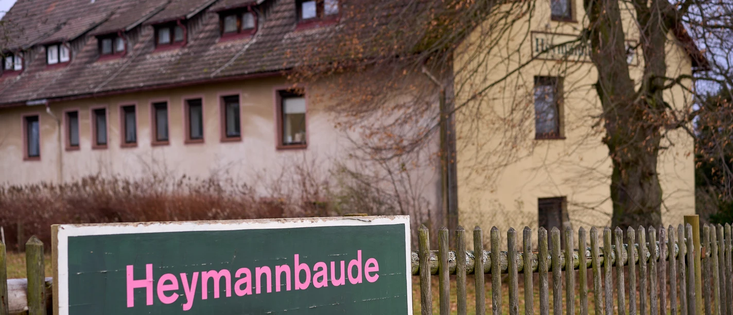 Heymannbaude in Kleinhennersdorf Wegweiser "Heymannbaude" mit Pfeil nach rechts, dahinter gelbes Gebäude und Zaun.