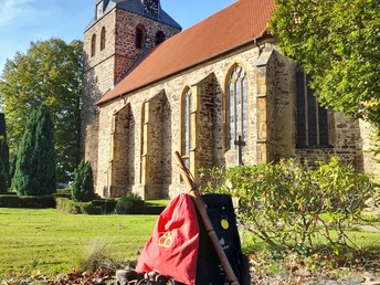 Gehlenbecker St. Nikolauskirche Pilgerausrüstung vor einer historischen Feldsteinkirche mit Turm und sonnigem Grünbereich