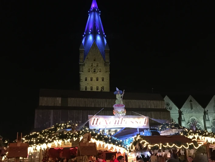 Weihnachtsmarkt bei Nacht: Menschen vor beleuchtetem Stand, beleuchteter Turm im Hintergrund.