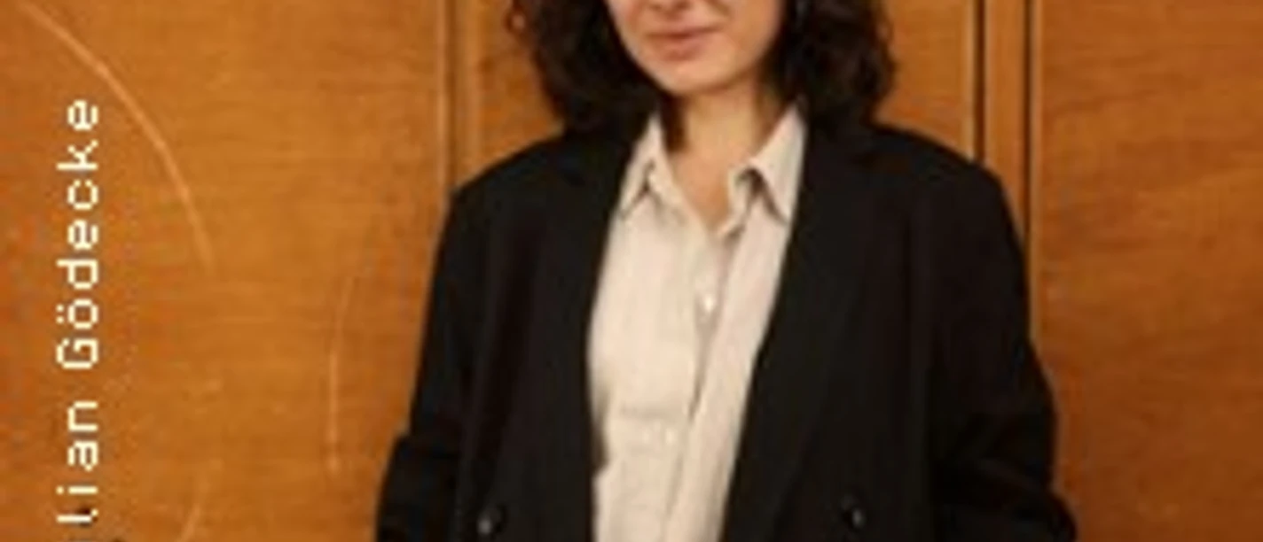 Person mit dunklem Haar im langen Mantel vor einer holzfarbenen Wand stehend.