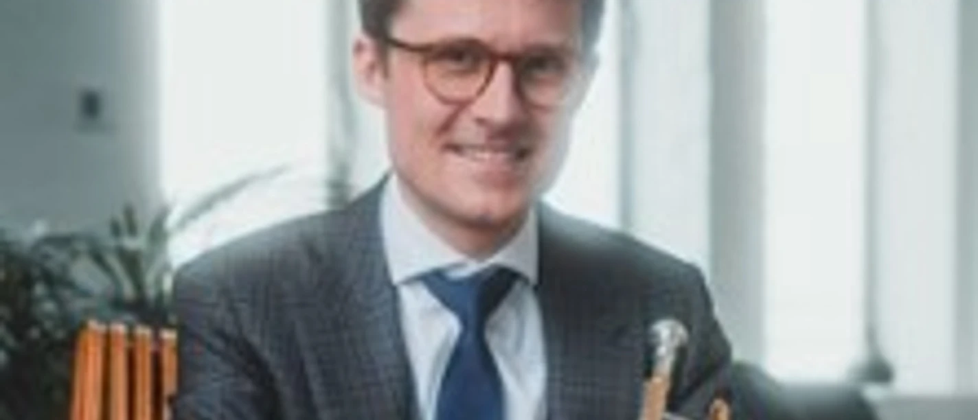 Ein lächelnder Mann im Anzug hält eine Trompete in einem hellen Raum mit hohen Fenstern.