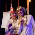 The Ransohoff case - women in carnival Zwei verkleidete Darstellerinnen auf einer Bühne vor einem Flipchart in bunter Beleuchtung.Two disguised actresses on a stage in front of a flipchart in colorful lighting.