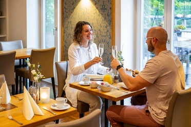 Espelkamp Schloss Benkhausen Zwei Personen stoßen beim Frühstück in einem hellen Raum an einem gedeckten Tisch an
