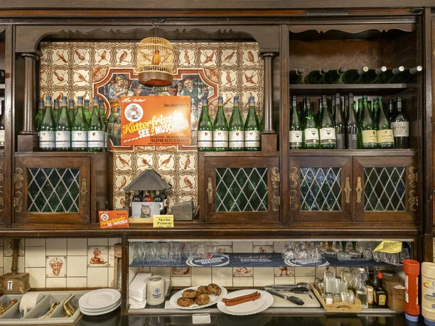 Weinhaus Vogel Holztheke mit Fliesenwand, Weinflaschen in Regalen und bereitgestellten Speisen.