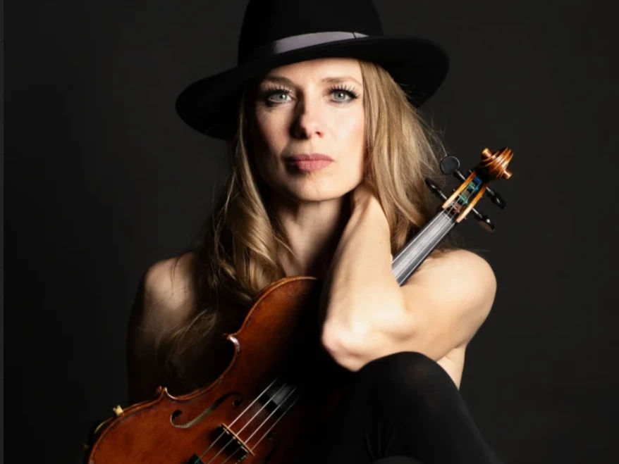 Meta Hüper Frau mit schwarzem Hut hält eine Violine vor dunklem Hintergrund
