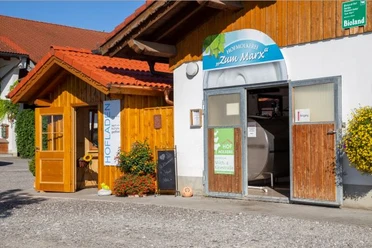 Hofmolkerei Marx Holzladen mit Blumen und offene Molkerei-Tür auf einem hellen Hof. Der Hof ist viel besucht.