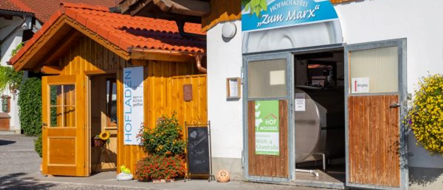Hofmolkerei Marx Holzladen mit Blumen und offene Molkerei-Tür auf einem hellen Hof. Der Hof ist viel besucht.