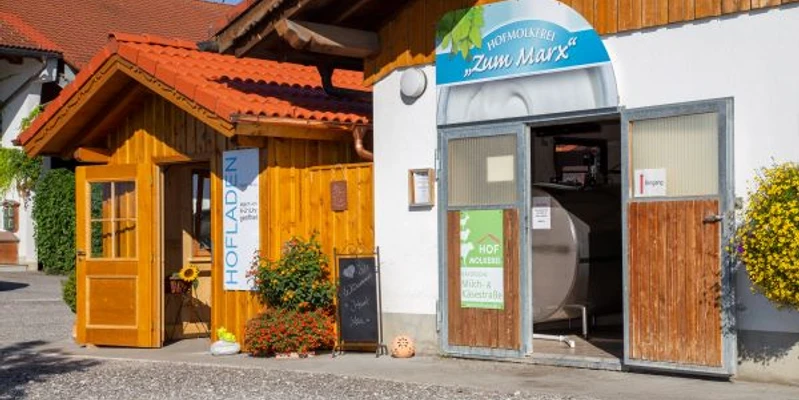Hofmolkerei Marx Holzladen mit Blumen und offene Molkerei-Tür auf einem hellen Hof. Der Hof ist viel besucht.