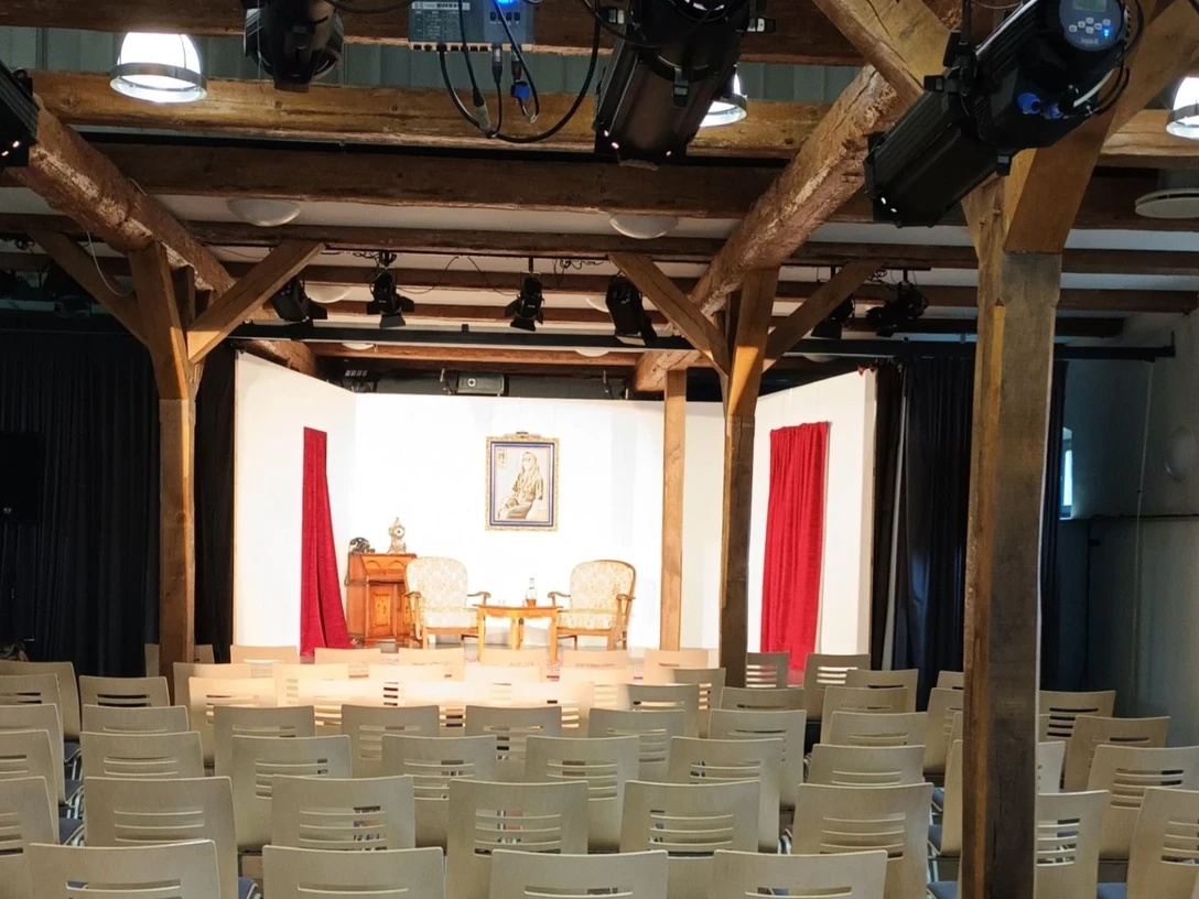 Pferdestall_innen_HD_16zu9.jpg Bühnenraum im Pferdestall mit Holzbalken, Stuhlreihen und Bühne mit rotem Vorhang und Scheinwerfern.