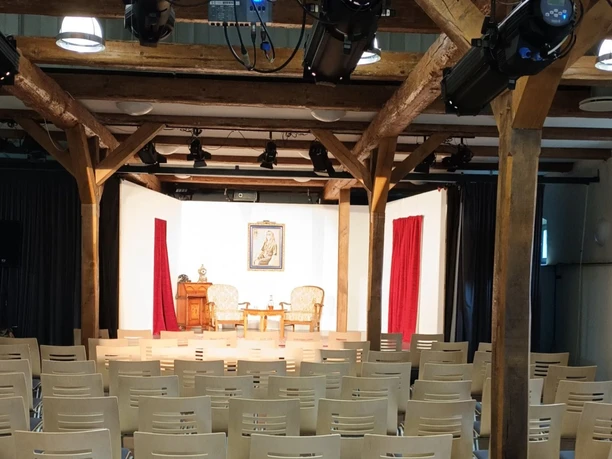 Pferdestall_innen_HD_16zu9.jpg Bühnenraum im Pferdestall mit Holzbalken, Stuhlreihen und Bühne mit rotem Vorhang und Scheinwerfern.