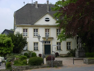 Altes Amtshaus - Rönsahl Blick auf das Bürgerhaus. Davor der Platz mit dem Denkmal und einigen Zierbüschen. Rechts und links flankieren Bäume den Platz.